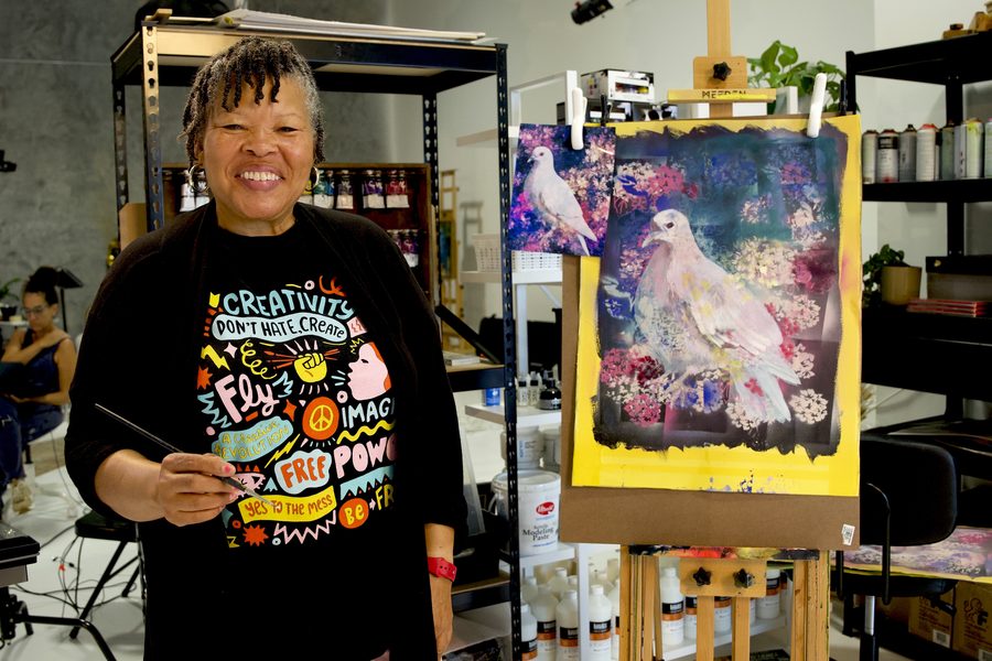 Artist laughing while holding brushes beside her colorful painting
