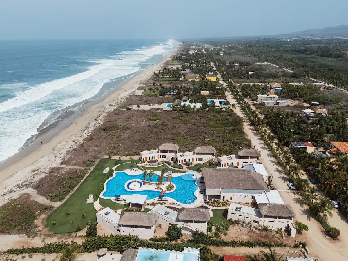 An aerial view along the Puerto Escondido coast: open ocean and breaking surf on the left, a long empty beach running into the distance, and the retreat property with its blue pool and thatched roofs set back from the sand