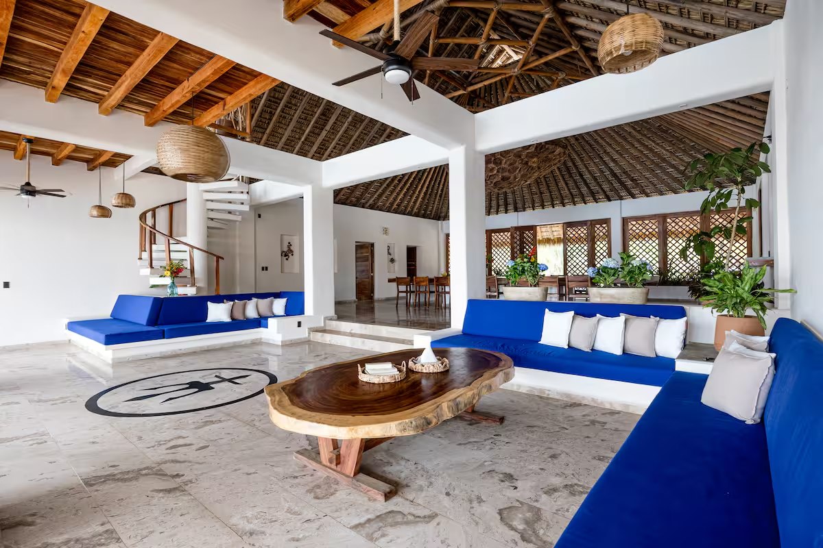 A large open-sided lounge with royal blue built-in sofas, a live-edge coffee table, a wood and thatch ceiling and a spiral stair