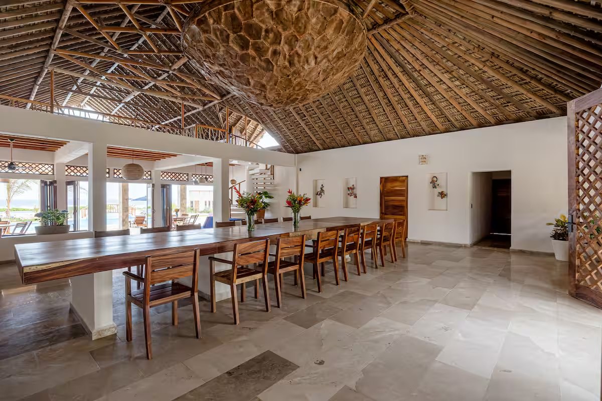 A large thatched dining hall with one long dark wood table lined with wooden chairs, flowers down the middle and a pale stone floor