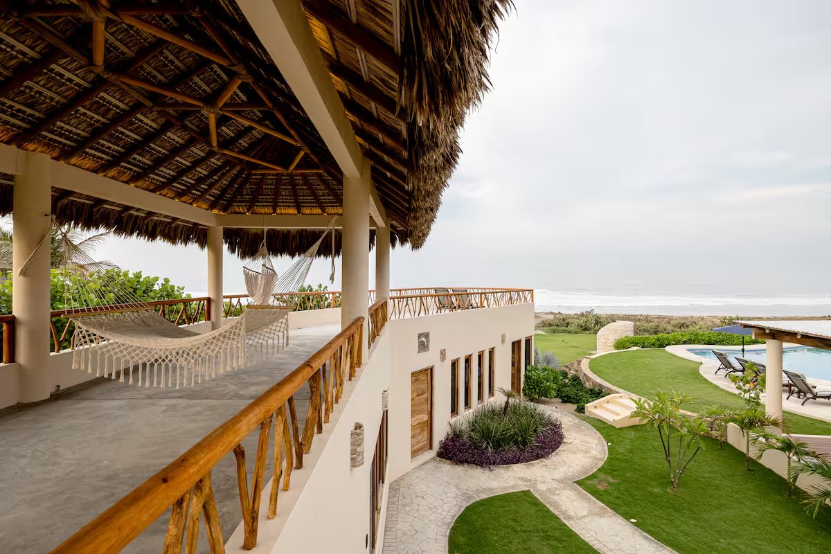 An upper walkway under a thatched roof with white hammocks slung along it, looking down over the lawn, the pool and the ocean