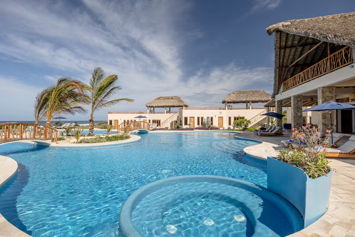 A wide daytime view of the pool complex with palms, umbrellas and thatched buildings around it