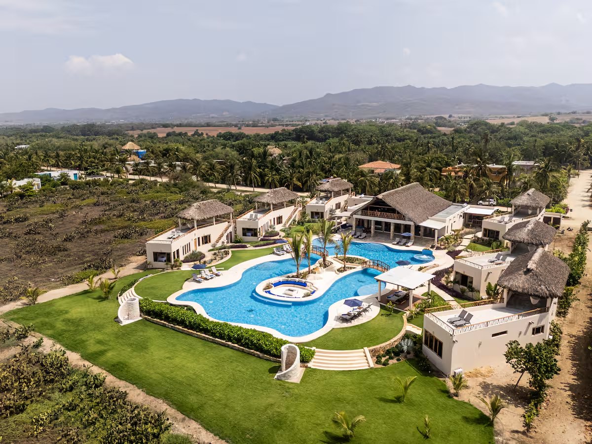 An aerial view of the property looking inland: a curved blue pool, a lawn, thatched-roof buildings and palms, with farmland and mountains behind