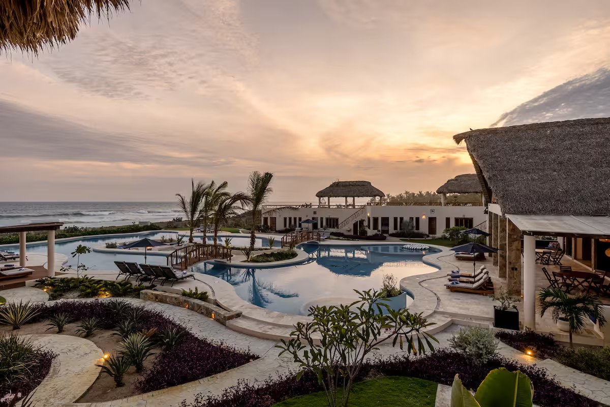 An aerial view of the retreat house at sunset: the curved pool lit from below, thatched palapas, palms, and the Pacific going orange behind them
