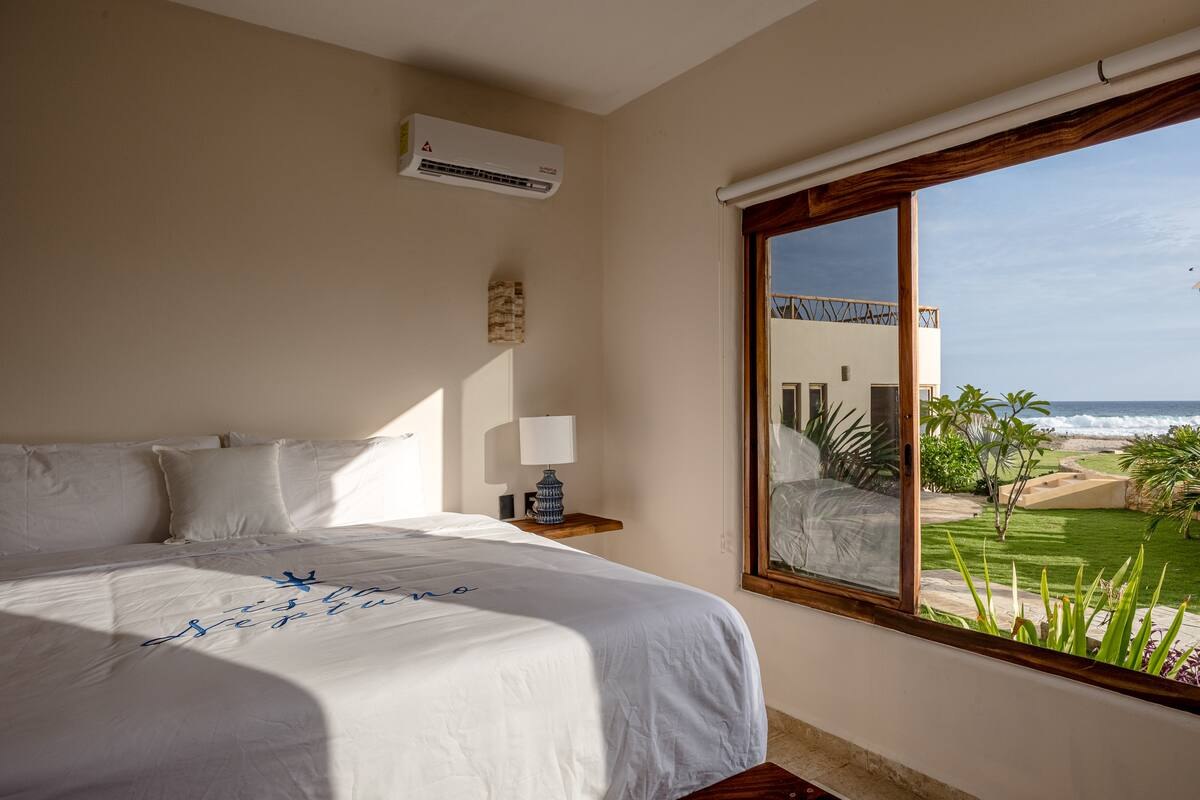 A guest room with a bed and a large window looking straight out to a lawn and the breaking surf