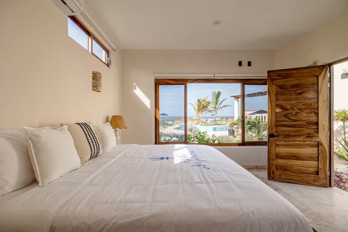 A guest room with a king bed and a window onto palms and a pool, with a carved wooden door standing open