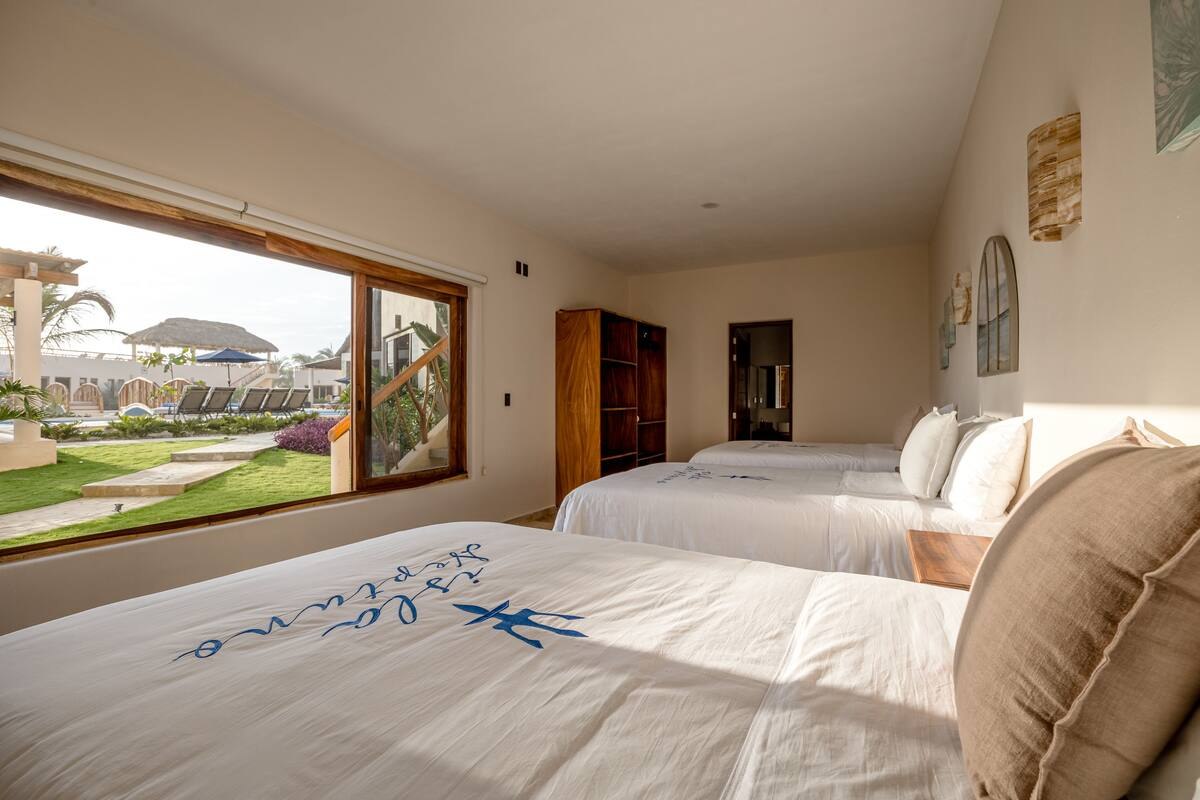 A guest room with two double beds in embroidered white linen and a picture window onto the lawn, pool and thatched buildings