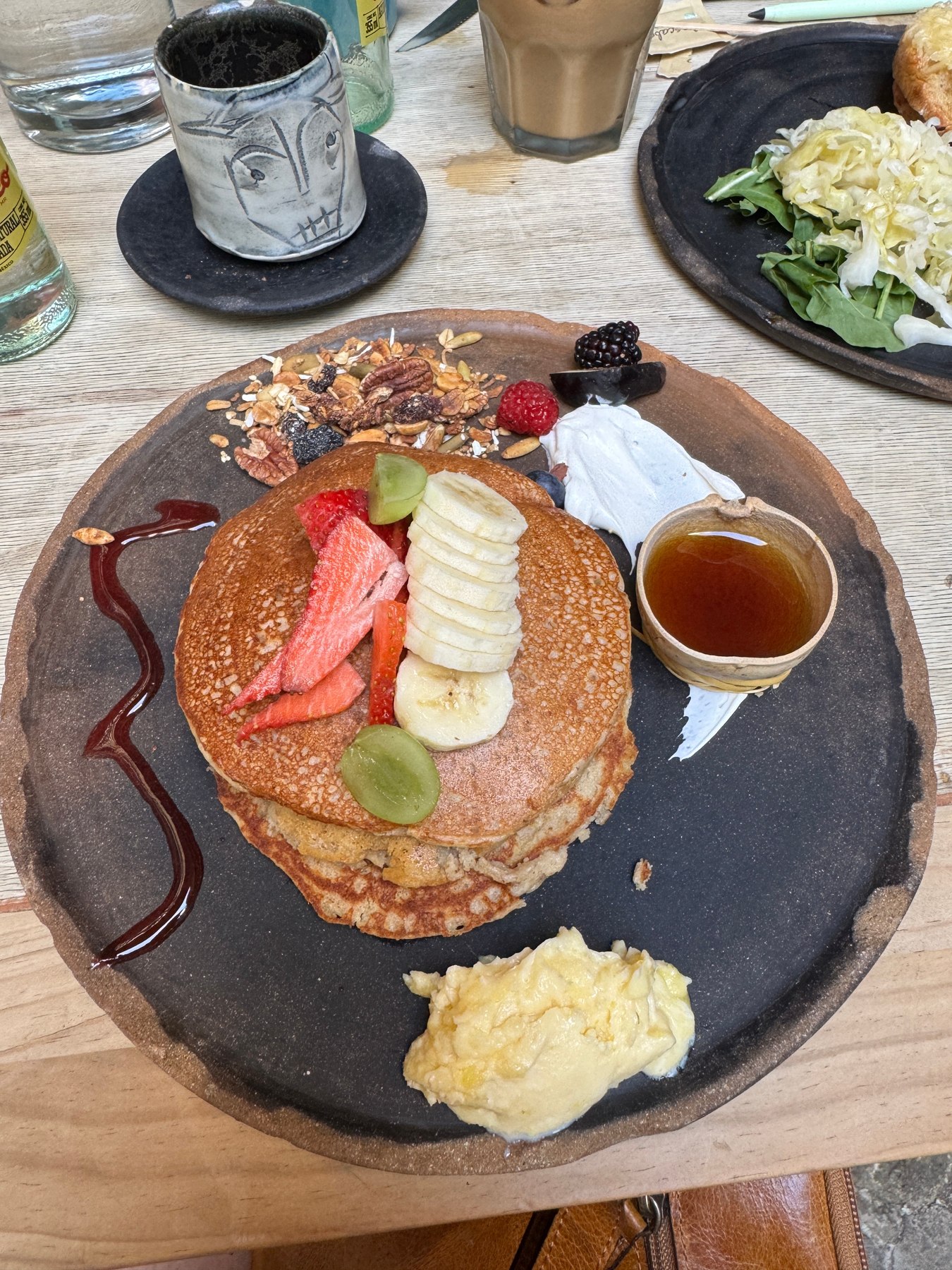 Breakfast on a dark ceramic plate: pancakes with strawberry, banana and granola, a jug of syrup, scrambled eggs and a hand-painted coffee cup