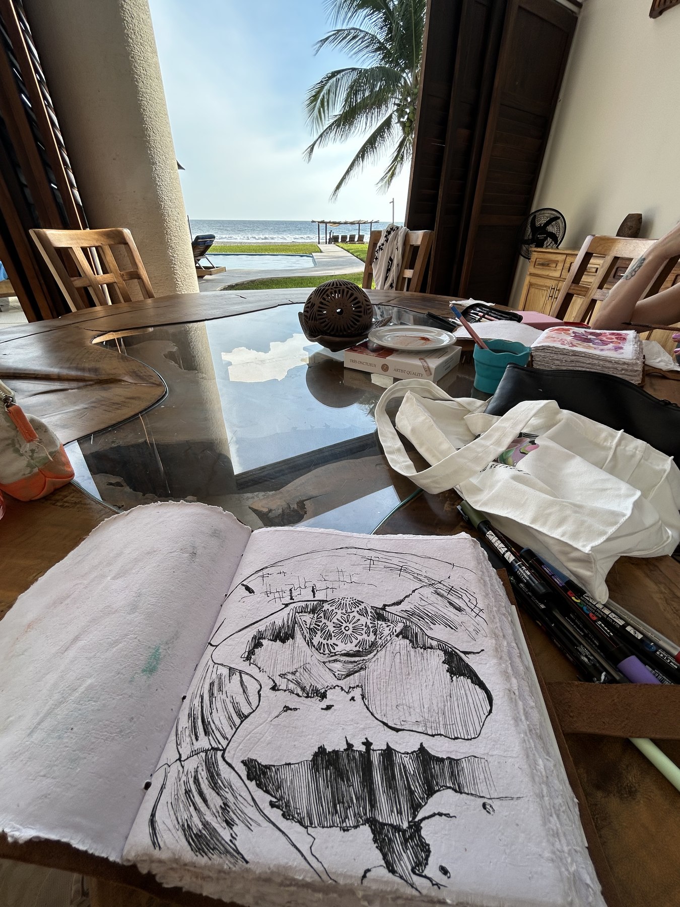 An open sketchbook with a black ink drawing on handmade paper, resting on a glass table beside a canvas tote and markers, with doors open to palms and the ocean