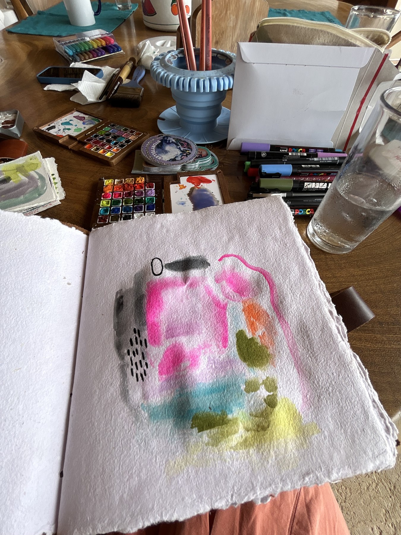 An open sketchbook of deckle-edged handmade paper showing a loose pink, grey and olive watercolor page, with pan palettes, markers, brushes and a water glass on a wooden table