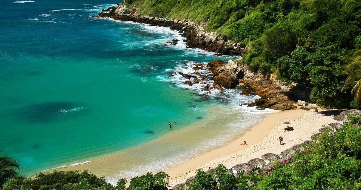 An aerial view of a small turquoise cove near Puerto Escondido: pale sand, green headlands on both sides, and a few swimmers in clear water