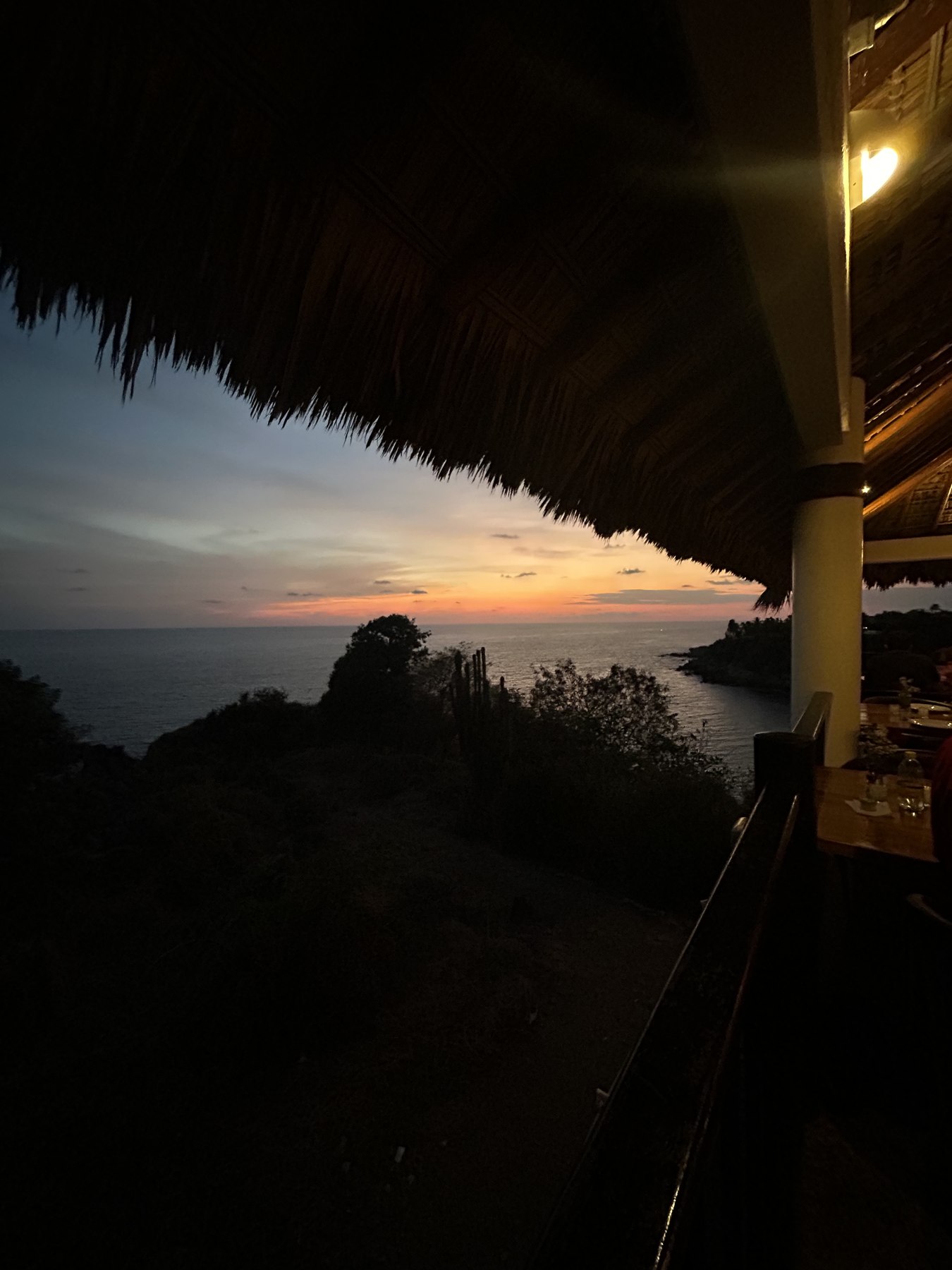 Dusk seen from a thatched cliffside restaurant looking over a cove, with set tables at the right of the frame