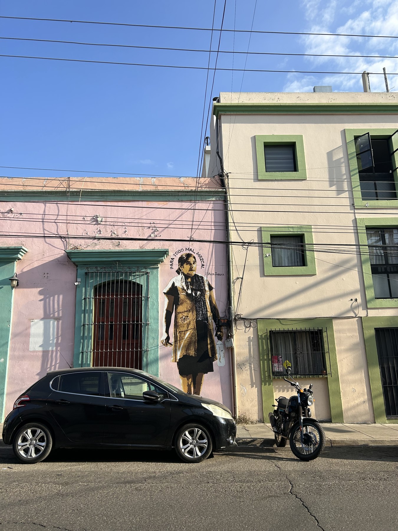 Pink and pale green colonial facades in Oaxaca City carrying a large painted mural of a woman in traditional dress, with a car and a motorcycle parked below