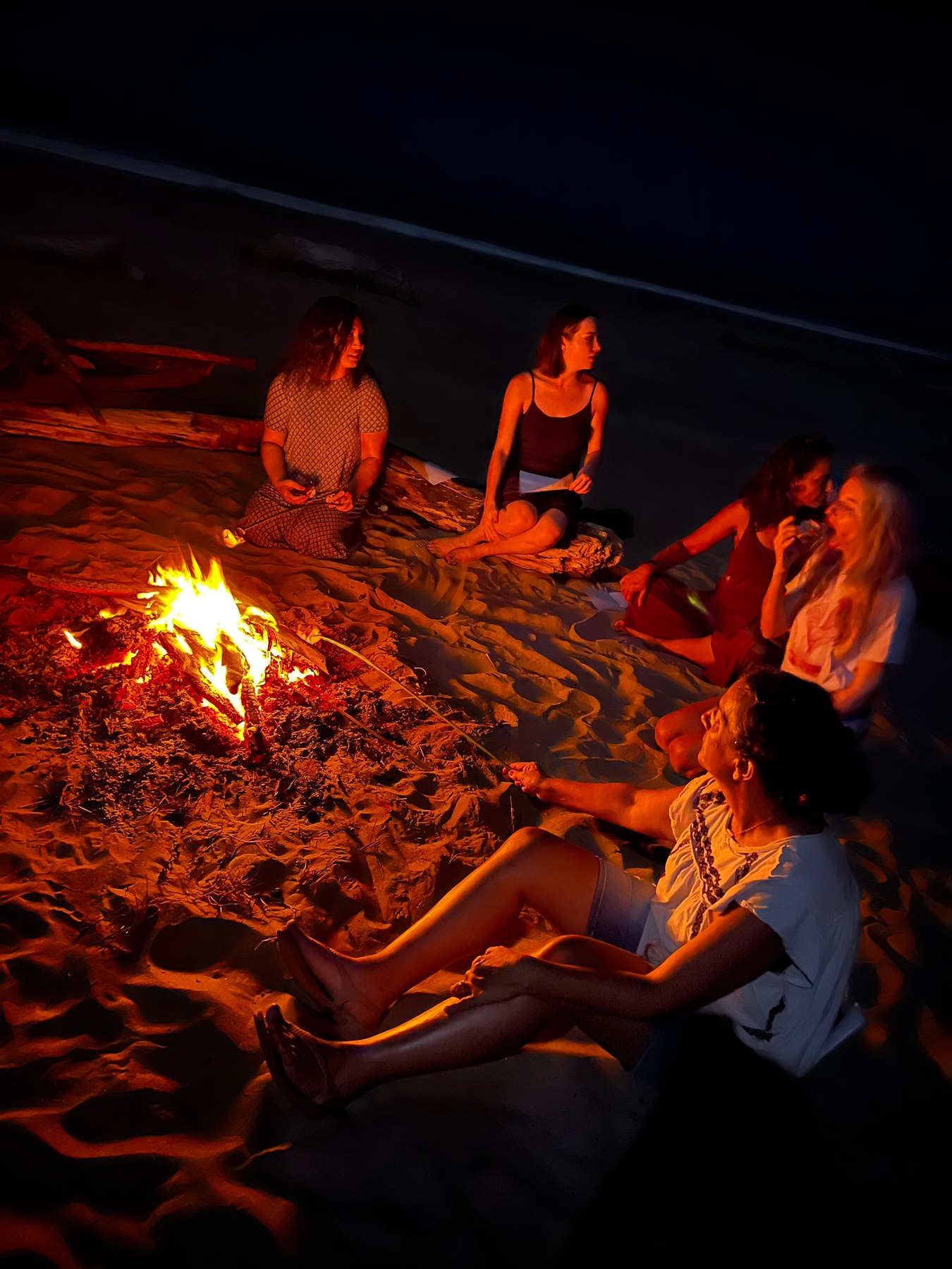 Five women sitting on the sand around a bonfire at night, lit by the firelight, talking and laughing