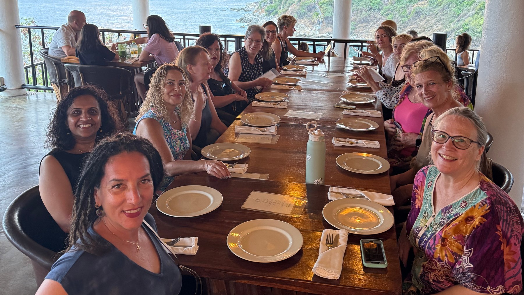 A group of women seated down both sides of one long wooden table on an open-air terrace, most turned toward the camera and smiling, with the ocean and a headland behind them