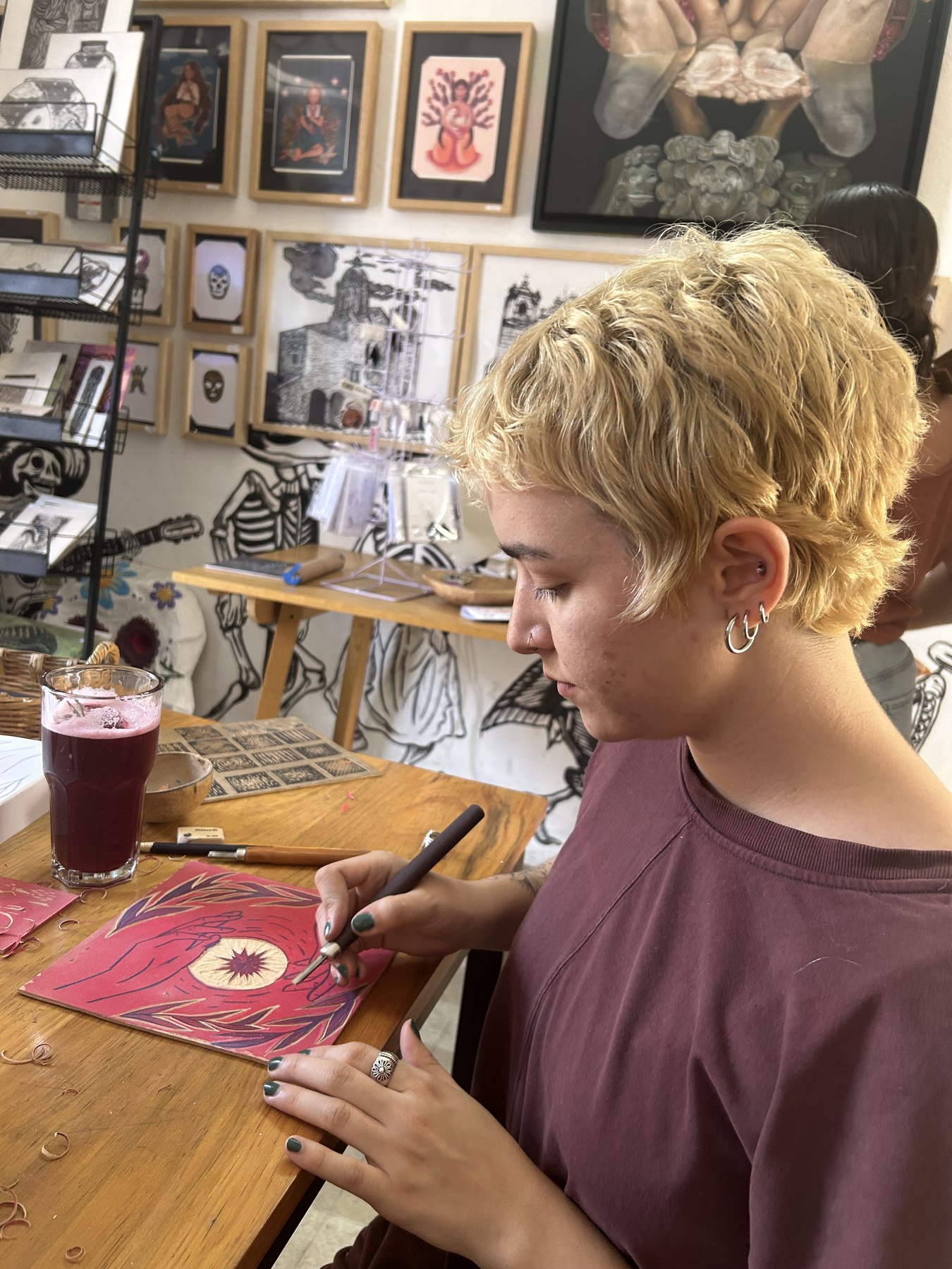 A person in profile carving a pink-inked block with a gouge, the design cut through the ink in white lines, wood shavings curled on the table, framed prints and skeleton artwork on the walls behind