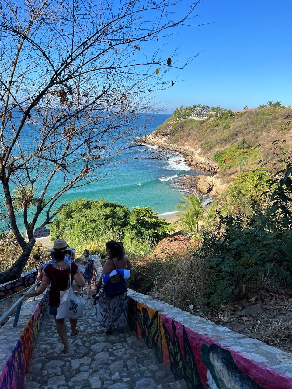 Women in sun hats carrying tote bags, walking down a long mosaic-tiled stairway toward a turquoise cove, with a headland and houses across the water