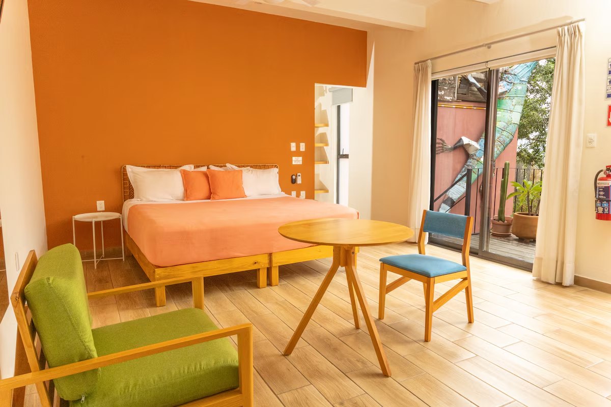 A room with a burnt-orange accent wall, a coral bedspread, a green armchair, a small round table and a sliding door onto a terrace