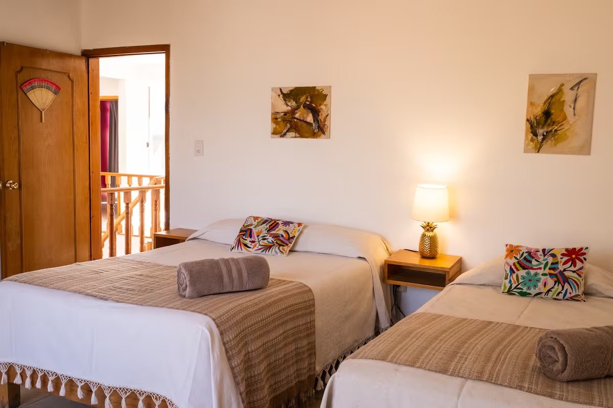 A twin room with two beds under woven blankets, Otomi embroidered cushions, a pineapple lamp and small bird paintings on the wall